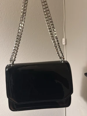 Svart axelväska  - Snygg svart axelväska i glansigt lackmaterial med dubbla silverfärgade kedjeremmar. Väskan har ett stilrent, fyrkantigt format och flera fack inuti, inklusive ett med dragkedja. 