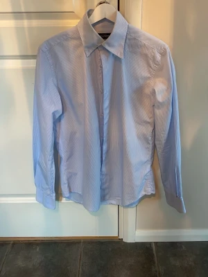 Ljusblå randig skjorta med knappar - Säljer en stilren ljusblå skjorta med tunna vita ränder och klassisk krage. Skjortan har långa ärmar och knäppning framtill. Perfekt till jeans eller chinos för en clean look.