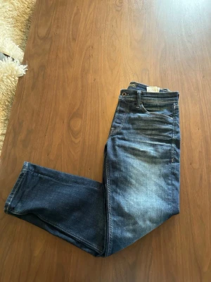 Blå raka jeans från Jack & Jones - Säljer ett par klassiska blå jeans från Nudie Jeans med rak passform och fem fickor. Använda 4 gånger tidigare. Jeansen har en snygg tvätt med lätt slitning framtill och detaljer i kontrastsömmar. Tillverkade i bomull med läderpatch bak i midjan.