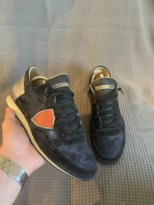 Philippe Model  - Snygga svarta sneakers från Philippe Model med mörkblått camouflagemönster och detaljer i mocka. Skorna har en röd logotyp på sidan och vita sulor. Perfekta för dig som vill ha en unik och trendig look.