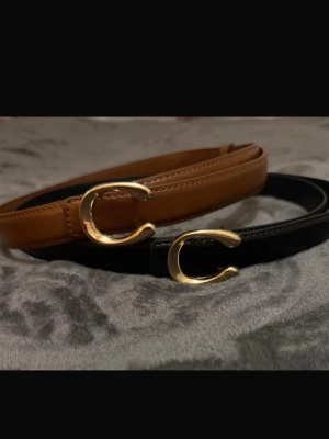 Coach bälten i svart och brunt skinn - Två stilrena bälten från Coach i svart och brunt skinn med ikoniska guldfärgade C-spännen. Perfekta för att lyfta vilken outfit som helst med en lyxig touch. Smal design som passar både till jeans och kj sopor.