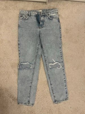 Ljusblå mom jeans med slitningar - Säljer ett par ljusblå mom jeans med hög midja och raka ben. Jeansen har slitna detaljer över båda knäna och klassiska fem fickor. Tillverkade i denim med vintagekänsla och har bälteshällor. Perfekta för en avslappnad och trendig look.
