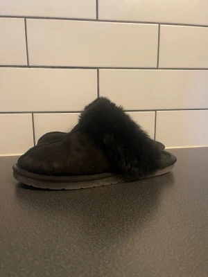 Svarta fluffiga slip-in tofflor - Mysiga svarta slip-in tofflor med mjuk och fluffig fuskpäls över vristen. Utsidan är i mockaimitation och sulan är platt och stabil. Perfekta för att hålla fötterna varma hemma. Klassisk och enkel design som passar till allt.