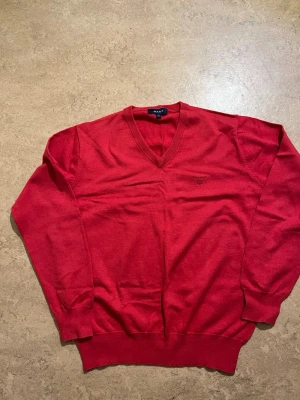 Röd V-ringad stickad tröja GANT - Snygg röd stickad tröja från GANT med V-ringning och diskret logga broderad på bröstet. Tröjan har långa ärmar och ribbade muddar vid ärmslut och nederkant. Perfekt för dig som vill ha en klassisk och stilren look.