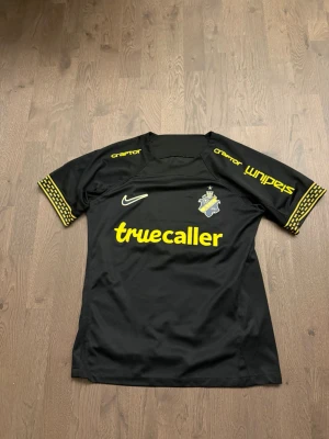 Svart AIK matchtröja Nike - Snygg svart AIK matchtröja från Nike med gula detaljer och tryck som truecaller och SVEA. Klubbmärket sitter på bröstet och ärmarna har mönstrade gula band. Perfekt för dig som vill stötta AIK på match eller träning.