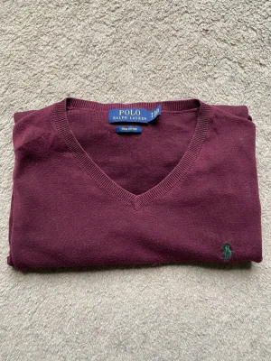 Vinröd stickad tröja Polo Ralph Lauren - Snygg vinröd stickad tröja från Polo Ralph Lauren i mjuk pima bomull. Tröjan har V-ringning och den klassiska gröna loggan broderad på bröstet. Perfekt för dig som gillar stilrena och tidlösa plagg. Vill bli av med den då den bara tar plats så säljer billigt.