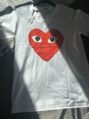 Vit Comme des Garçons Play t-shirt - Säljer en vit t-shirt från Comme des Garçons Play med det ikoniska röda hjärtat med ögon på bröstet. T-shirten har korta ärmar och är gjord i mjuk bomull. Perfekt för dig som gillar streetwear och vill ha något stilrent men ändå lekfullt.