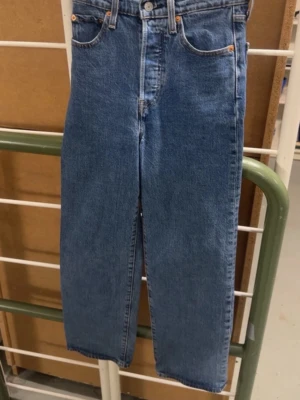 Blå Levi's raka jeans - Klassiska blå Levi's jeans med rak passform och hög midja. Jeansen har fem fickor, knappgylf och tydliga sömmar. Tillverkade i slitstark denim med den ikoniska Levi's-lappen bak i midjan. Perfekta för en avslappnad och tidlös stil.