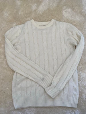 Vit kabelstickad tröja - Kabelstickad tröja i mjuk vit färg med rund halsringning och ribbade muddar vid ärmslut och nederkant. Klassisk design med vertikala flätmönster som ger en tidlös look. Perfekt för lager på lager under kyliga dagar.