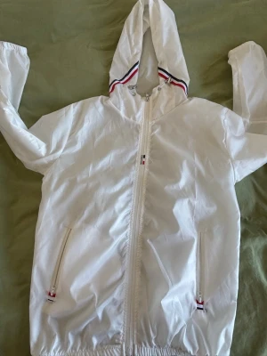 Vit windbreaker från Moncler - Snygg vit vindjacka från Moncler med huva och dragkedja framtill. Jackan har två fickor med dragkedja och Moncler-logga på ärmen. Detaljer i rött, vitt och blått vid huva och fickor. Tillverkad i lätt, glansigt material som passar perfekt till streetwear.