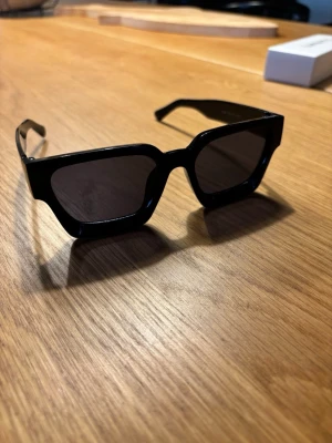 Chimi 04 svarta liknade - Säljer ett par svarta solglasögon med breda, kantiga bågar och mörka glas. De har en modern och edgy look som verkligen sticker ut. Perfekta för dig som vill ha en statement-accessoar till sommaren.