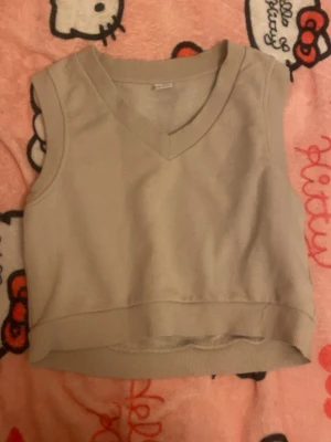 Beige ärmlös väst från Lindex - Säljer en beige, ärmlös väst från Lindex med v-ringad hals och bred ribbad kant nedtill. Perfekt att slänga över en skjorta eller t-shirt för en enkel och clean look. Tillverkad i mjuk bomull för skön känsla.
