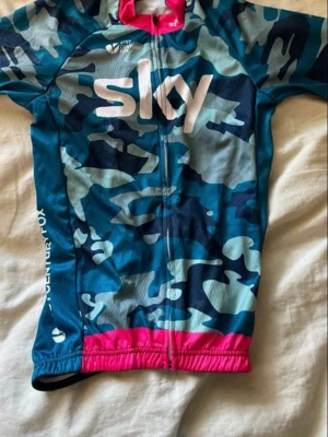 Blå camo cykeltröja och byxor från Sky - Säljer en blå camouflagemönstrad cykeltröja och byxor från Sky med dragkedja framtill och rosa detaljer vid nederkanten. Tröjan har korta ärmar, tre fickor bak och är tillverkad i ett lätt, ventilerande material. Perfekt för cykelturen!