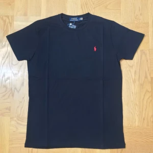 Svart t-shirt från Polo Ralph Lauren - Klassisk svart t-shirt från Polo Ralph Lauren med röd broderad logga på bröstet. T-shirten har rund halsringning och korta ärmar. Tillverkad i mjuk bomull för en skön känsla och normal passform.