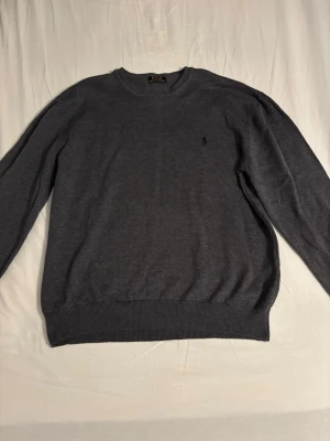 Mörkgrå tröja från Polo Ralph Lauren - Mörkgrå rundhalsad tröja från Polo Ralph Lauren med diskret broderad logga på bröstet. Tröjan har långa ärmar och ribbade muddar vid ärmslut och nederkant. Perfekt för en stilren och enkel look.