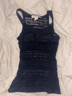 Mörkblå spetslinnetopp med knappar - Säljer en mörkblå linnetopp från HM med spetsdetaljer och tre små knappar framtill. Toppen är ärmlös och har ett figurnära snitt. Perfekt att styla lager på lager eller som statement-piece till jeans.