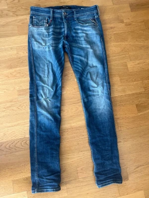 Blå slitna jeans från Replay - Säljer ett par blå jeans från Replay med slitningar och distressed detaljer på låret och knät. Klassisk femficksmodell med smal passform och snygga kontrastsömmar. Materialet är bomull med lite stretch för extra komfort.