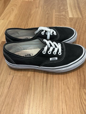 Svarta Vans sneakers med vita detaljer - Klassiska svarta Vans sneakers med vit sula och vita skosnören. Skorna har låg profil och tydliga vita sömmar som kontrast. Ovandelen är i canvasmaterial och har Vans-logga på sidan. Perfekta för en avslappnad och stilren look.