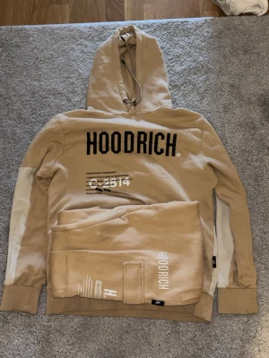 Beige tracksuit från Hoodrich - Säljer en beige hoodie från Hoodrich med svart logga och tryck framtill samt coolt kartmönster och text på ryggen. Hoodien har huva med dragsko och breda muddar. Materialet är mjukt och skönt, perfekt för en avslappnad streetwear-look. Byxorna är också storlek M.