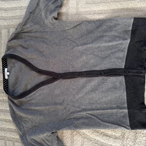 Grå cardigan med armbågslappar - Snygg grå cardigan med mörkare knäppning och ribbade muddar. Tröjan har dekorativa armbågslappar i matchande färg och knappar längs hela framsidan. Perfekt för dig som gillar en stilren och enkel look.