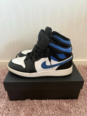 Air Jordan 1 mid svart/blå/vit - Snygga Air Jordan 1 High sneakers i svart, blått och vitt läder. Klassisk siluett med högt skaft, blå Swoosh och ikoniska Air Jordan-loggan på sidan. Skorna har svart snörning och vit mellansula för en clean look.