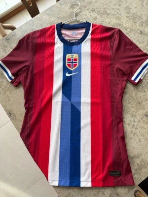 Norge fotbollströja Nike röd/blå - Säljer en officiell Norge fotbollströja från Nike med vertikala ränder i rött, vitt och blått. Tröjan har Norges landslagsemblem på bröstet och klassisk Nike-logga. Tillverkad i lätt och ventilerande material, perfekt för träning eller match.