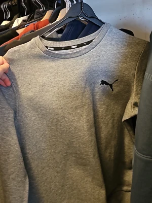 Grå sweatshirt från Puma - Klassisk grå sweatshirt från Puma med rund halsringning och svart Puma-logga på bröstet. Tröjan har en enkel och stilren design, perfekt för en avslappnad look. Tillverkad i mjukt material för extra komfort.