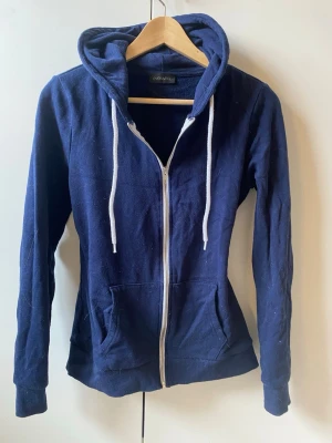 Mörkblå zip hoodie med vita snören - Mörkblå hoodie med dragkedja framtill, vita snören i huvan och två fickor på magen. Tillverkad i mjukt bomullsmaterial och har ribbade muddar vid ärmslut och nederkant. Perfekt för en avslappnad och stilren look. riktigt bra skick då den inte har vart använd många ggr!! pass formen är ”fitted” asså ganska tajt men inte tajt tajt 