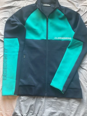 J.Lindeberg zip -  Snygg golf cardigan från J.Lindeberg i marinblått och grönt med dragkedja framtill. Märket syns tydligt på bröstet och längs ena ärmen. Den har hög krage, två fickor med dragkedja och sportig design. Perfekt för dig som gillar stilrena och moderna plagg nu till golfsäsongen eller bara vardagsplagg. Den är aldrig använd då den är för liten för mig. 