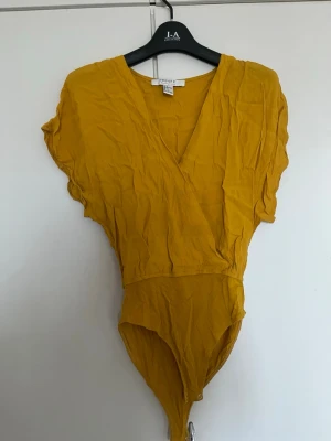 Gul body från Forever 21 - Snygg gul body från Forever 21 med korta ärmar och omlott-ringning. Tillverkad i ett lätt, mjukt tyg som ger en avslappnad look. Perfekt att styla med jeans eller kjol för en trendig outfit.
