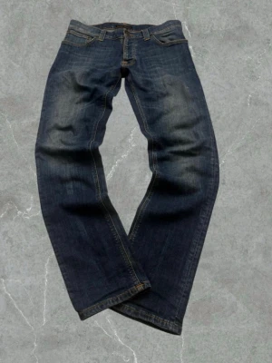 Mörkblå jeans från Nudie Jeans - Säljer ett par klassiska mörkblå jeans från Nudie Jeans med snygg slitning och raka ben. Jeansen har orangea sömmar och är tillverkade i slitstark denim. Perfekta för dig som gillar en tidlös och avslappnad stil.