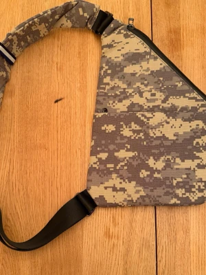 Camouflage axelväska från NIID - Säljer en snygg axelväska från NIID med digitalt camouflagemönster i beige, grått och grönt. Väskan har en trekantig form, justerbar rem och dragkedja upptill. Perfekt för dig som vill ha en unik och praktisk väska till vardags. Sprillans ny,aldrig använd! 