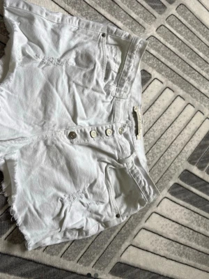Vita jeansshorts med knappar - Säljer ett par vita jeansshorts från One Teaspoon med råa kanter och slitna detaljer. Shortsen har hög midja, knappgylf och klassiska fem fickor. Perfekta för sommaren och ger en avslappnad look.