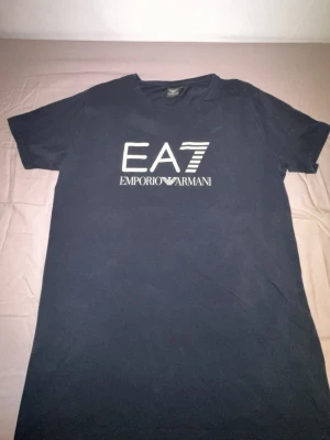 Mörkblå EA7 Emporio Armani t-shirt - Snygg mörkblå t-shirt från EA7 Emporio Armani med stort vitt tryck på bröstet. Klassisk rund hals och korta ärmar. Tillverkad i mjuk bomull för skön känsla och stilren look.