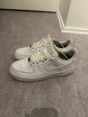 Nike Air Force 1  - Klassiska vita Nike Air Force 1 sneakers i lågt utförande. Skorna har en stilren design med perforerad tå, vita skosnören och den ikoniska swoosh-loggan på sidan. Tillverkade i läder med en robust gummisula för bra grepp. Perfekta för dig som gillar streetwear och enkel stil.