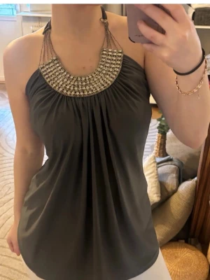 Vintage❤️ - Snygg mörkgrå halterneck topp med dekorativa kedjor och glittriga stenar runt halsringningen. Toppen har ett löst fall och är ärmlös, perfekt för att sticka ut med ett statement-plagg. Passar till både jeans och kjol för en trendig look.