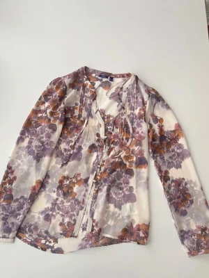 Blommig blus med knappar - Snygg blus med blommigt mönster i lila, orange och vitt. Blusen har långa ärmar, knappar framtill och dekorativa veck vid axlarna. Perfekt för en trendig och feminin look.