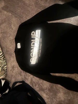 Svart sweatshirt från Dondup - Svart sweatshirt från Dondup med stor vit logga och texten 'Unconventional Clothing Established 1999' på bröstet. Tillverkad i Italien. Klassisk rak passform och rund halsringning. Perfekt för en clean och stilren look. Storlek M ganska liten i storleken passar mer S 