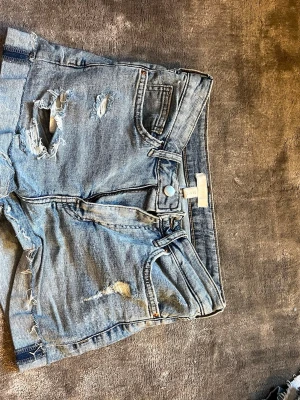 Blå slitna jeansshorts från H&M - Säljer ett par blå jeansshorts från H&M med trendiga slitningar och råa kanter. Shortsen har klassisk femficksdesign, knappgylf och är gjorda i denim. Perfekta för en avslappnad och cool stil i sommar.