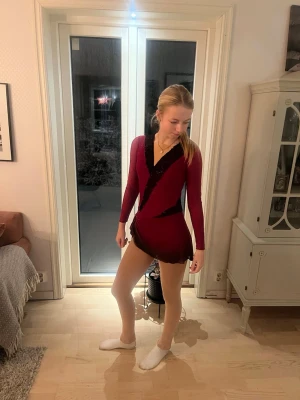 Vinröd konståkningsklänning - Snygg vinröd konståkningsklänning med långa ärmar och svarta diagonala detaljer. Klänningen har djup V-ringning och glittrande stenar på framsidan. Kjolen är kort och har vågig kant. Perfekt för träning eller tävling på isen.