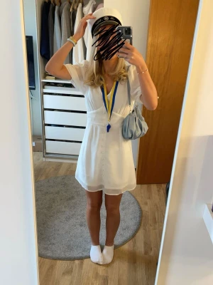 Studentkläning  - Säljer en söt vit kort klänning med v-ringning och knappar framtill. Klänningen har korta ärmar och lätt volang nedtill. Perfekt till student eller sommarens alla tillfällen. Enkel och stilren design som passar till mycket.
