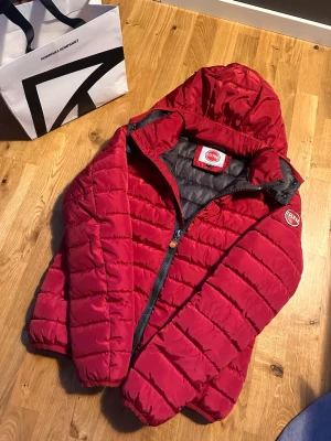 Colmar jacka - Snygg röd dunjacka från Colmar med huva och svart innerfoder. Jackan har dragkedja framtill, elastiska muddar och Colmar-logga på ärmen. Perfekt för kalla vinterdagar och riktigt skön att bära. Klassisk quiltad design som håller värmen bra.