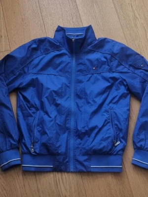 Blå vindjacka från Tommy Hilfiger - Snygg blå vindjacka från Tommy Hilfiger med dragkedja framtill, två fickor med dragkedja och diskret logga på bröstet. Jackan har ribbade muddar med vita detaljer och står upp i kragen. Tillverkad i lätt, glansigt material som passar perfekt till streetwear.