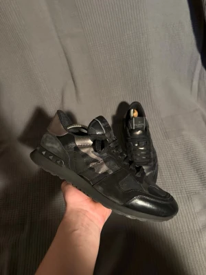 Valentino rockrunner - Snygga svarta sneakers från Valentino med diskret camomönster och detaljer i mocka och läder. Skorna har svarta snören, robust sula och coola nitar på hälen. Perfekta för dig som vill ha en exklusiv och trendig look.