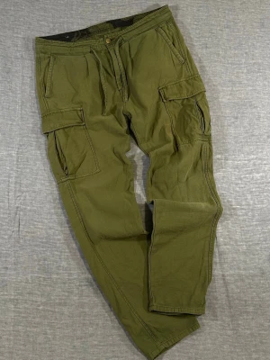 Scotch & Soda olive green cargo pants W33 L34 military style - Scotch & Soda cargo pants in olive green. Military-inspired design with straight tapered fit, durable 100% cotton, functional cargo pockets, and drawstring waist detail. Clean utility look perfect for workwear or streetwear.  Waist 46 cm, Rise 29 cm, Thigh 30 cm, Knee 22 cm, Foot 17 cm, Inseam 76 cm.