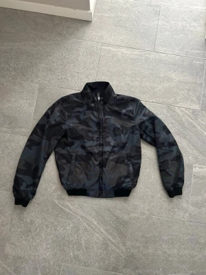 Svartblå bomberjacka med camomönster - Snygg bomberjacka i svart och mörkblått camomönster. Jackan har dragkedja framtill, ribbade muddar vid ärmslut och midja samt två sidofickor. Materialet är lätt och ger en sportig look. Perfekt för dig som vill ha en streetinspirerad stil.