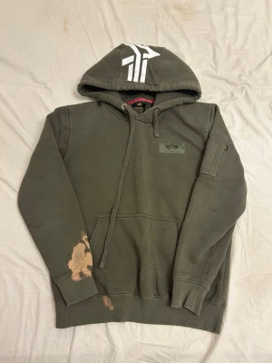 Grön hoodie från Alpha Industries - Snygg grön hoodie från Alpha Industries med stor vit logga på huvan och patch på bröstet. Klassisk känguruficka framtill, dragsko i huvan och ribbade muddar. Tillverkad i mjuk bomullsmix och har en unik detalj med ficka på ärmen.