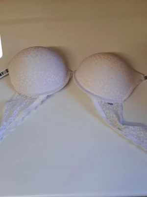 Vit spets-bh från Calvin Klein - Snygg vit bh från Calvin Klein med vadderade kupor och spetsdetaljer på sidorna. Axelbanden är justerbara och har logotypen tryckt längs hela bandet. Bh:n har en feminin och stilren design med mjuk spets och släta kupor.