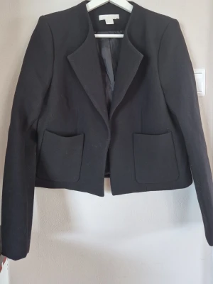 Kort svart kavaj med axelvadd fickor från H&M - Stilren svart kavaj från H&M med två stora framfickor och klassisk krage. Kavajen har en rak, något kortare passform och liten axelvadd. Materialet har snygg struktur. Perfekt för dig som vill ha en enkel men trendig look.
