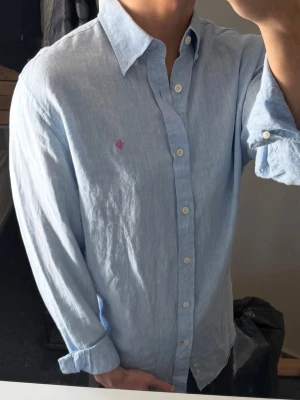 Ljusblå linneskjorta från Morris - Säljer en ljusblå skjorta från Morris i 100% linne med klassisk krage och knappar framtill. Skjortan har lång ärm och en liten rosa logga broderad på bröstet. Perfekt för en avslappnad men ändå stilren look. Lite missfärgad på insidan av kragen, annars bra skick